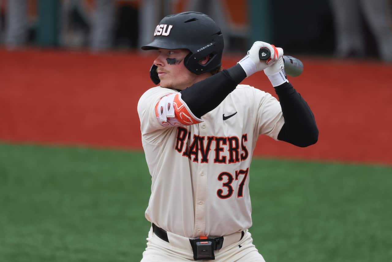 A man wearing a white baseball jersey and black helmet swings a bat back, ready to receive a pitch. 