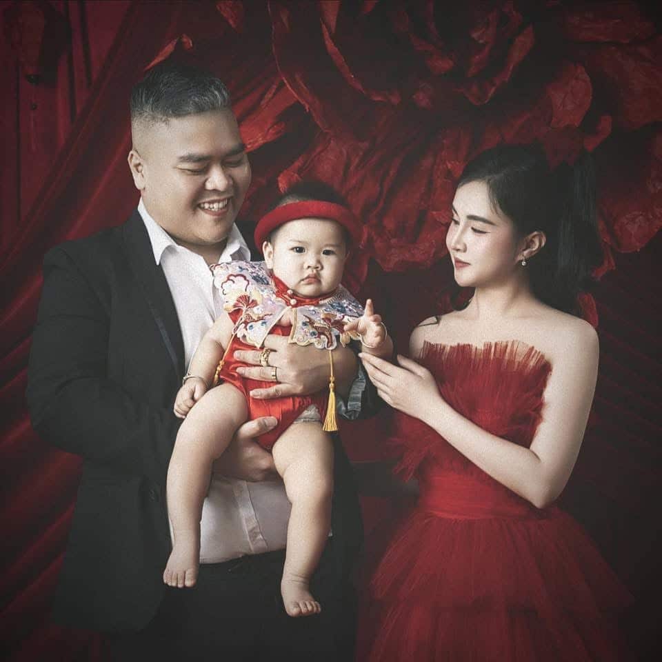 A man in a suit jacket holds a baby while a woman in a red dress stands at their side with her left hand touching the baby.