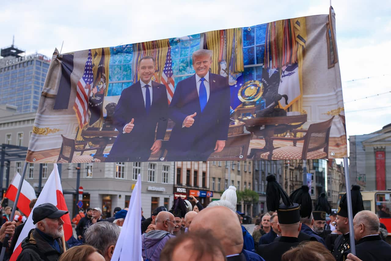 A banner featuring two men in blue suits, giving thumbs-up signs, is being held up by people at a rally.