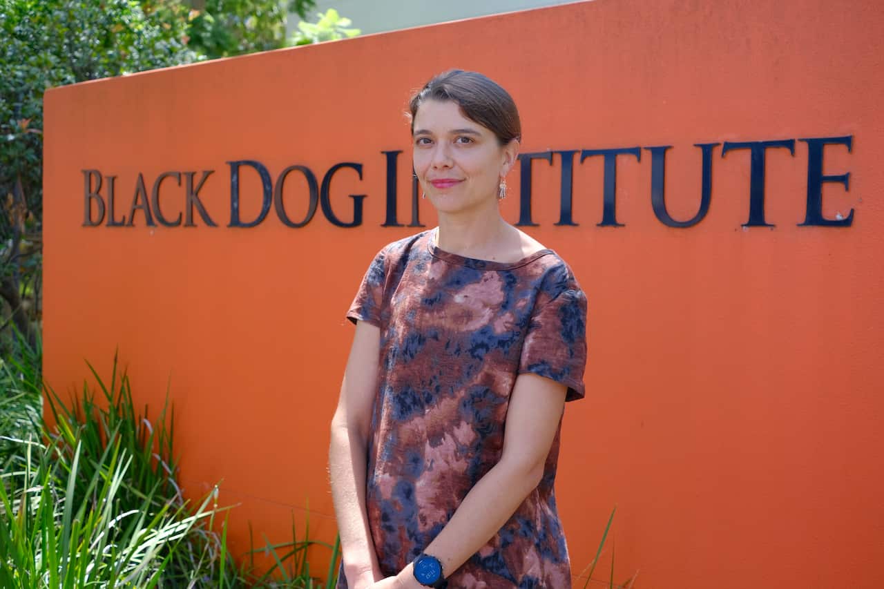 A woman stands in front of an ochre-coloured wall outside. The wall has the words Black Dog Institute it in black lettering