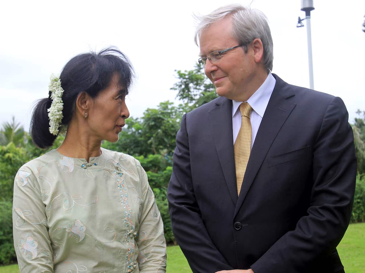 Aung San Suu Kyi, wearing a traditional light green blouse with flowers in her hair, stands outdoors looking toward Australian politician Kevin Rudd, who is dressed in a dark suit and gold tie.