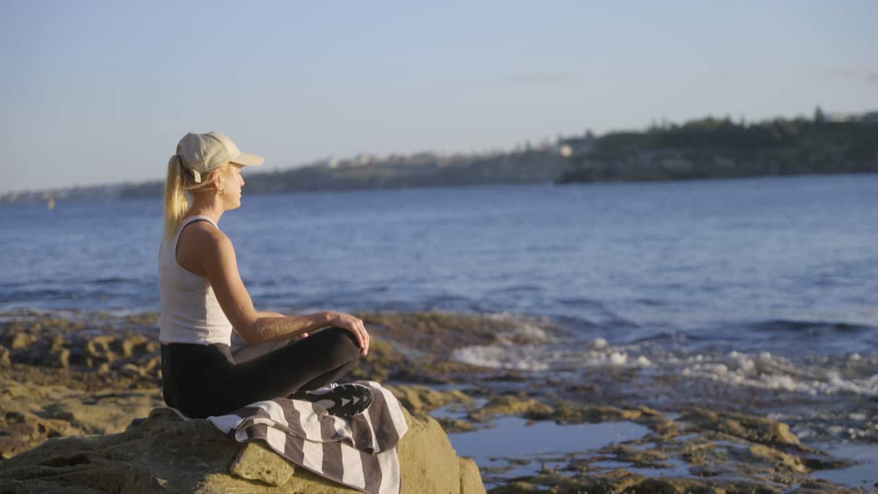 A woman sits on a rock facing the ocean with her legs crossed.