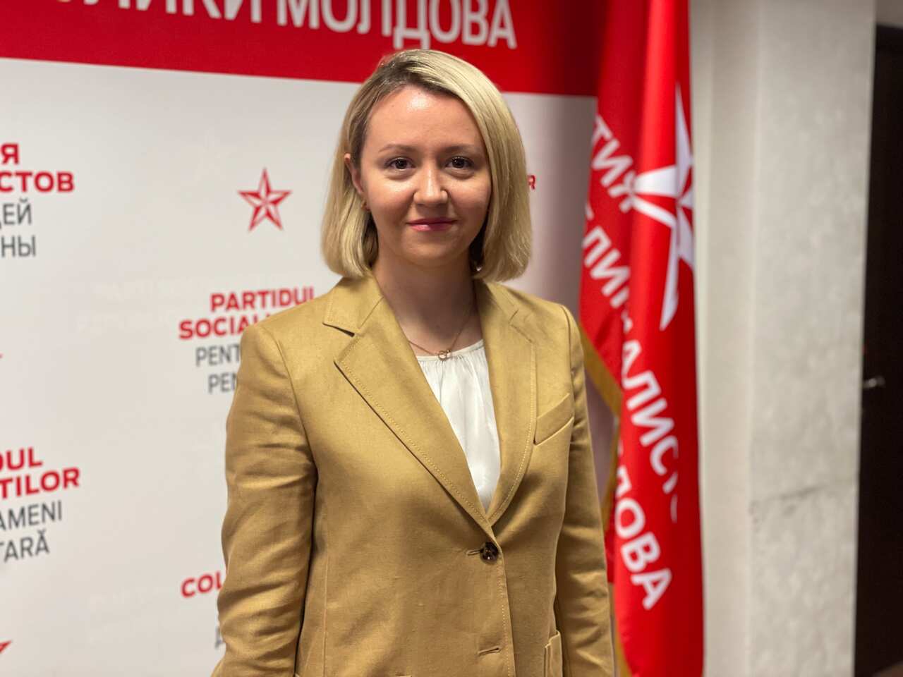 A young woman with blonde hair wearing a tan blazer standing in front of a red flag and poster. 