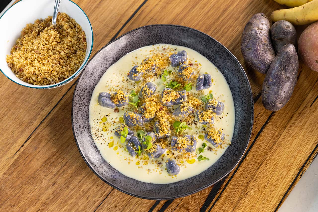 A wide silver-brown pottery bowl sits on a wooden surface. It holds purple gnocchi in a creamy sauce. A bowl of cooked breadcrumb topping sits alongside. Some has been sprinkled on the gnocchi, along with small herb leaves. 