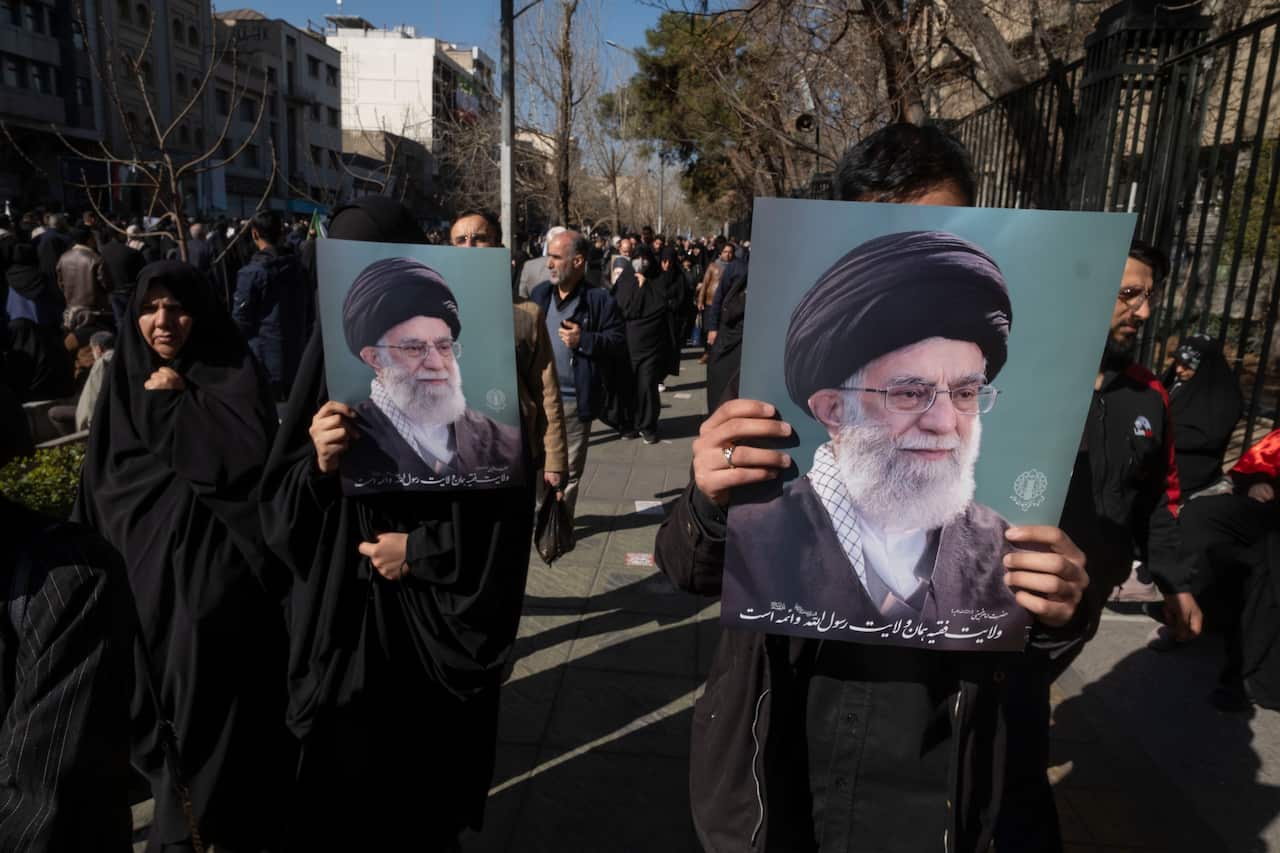 People in a crowded outdoor walkway carry large portraits of Iranian Supreme Leader Ali Khamenei during a public gathering.