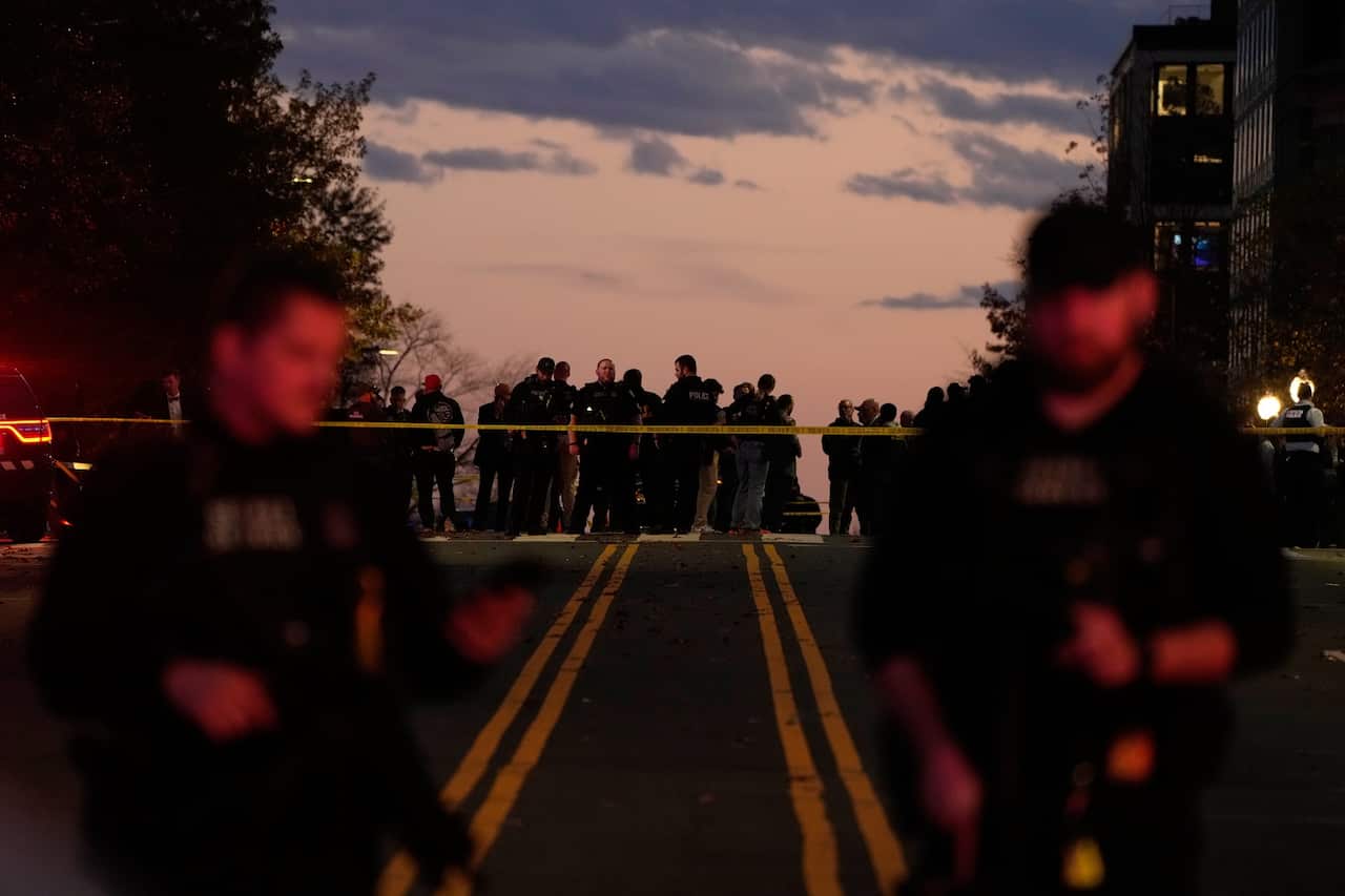 Armed personnel stand on a road in front of a tape that cordons off an area of the incident. A crowd is visible on the other side of the tape.