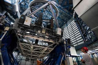 A man wearing a red helmet and grey shirt looks up at a large telescope inside an industrial centre.