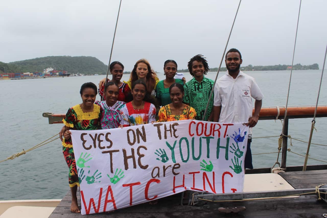 A group of young people hold up a sign on a boat. The sign reads Eyes on the court, the youth are watching.