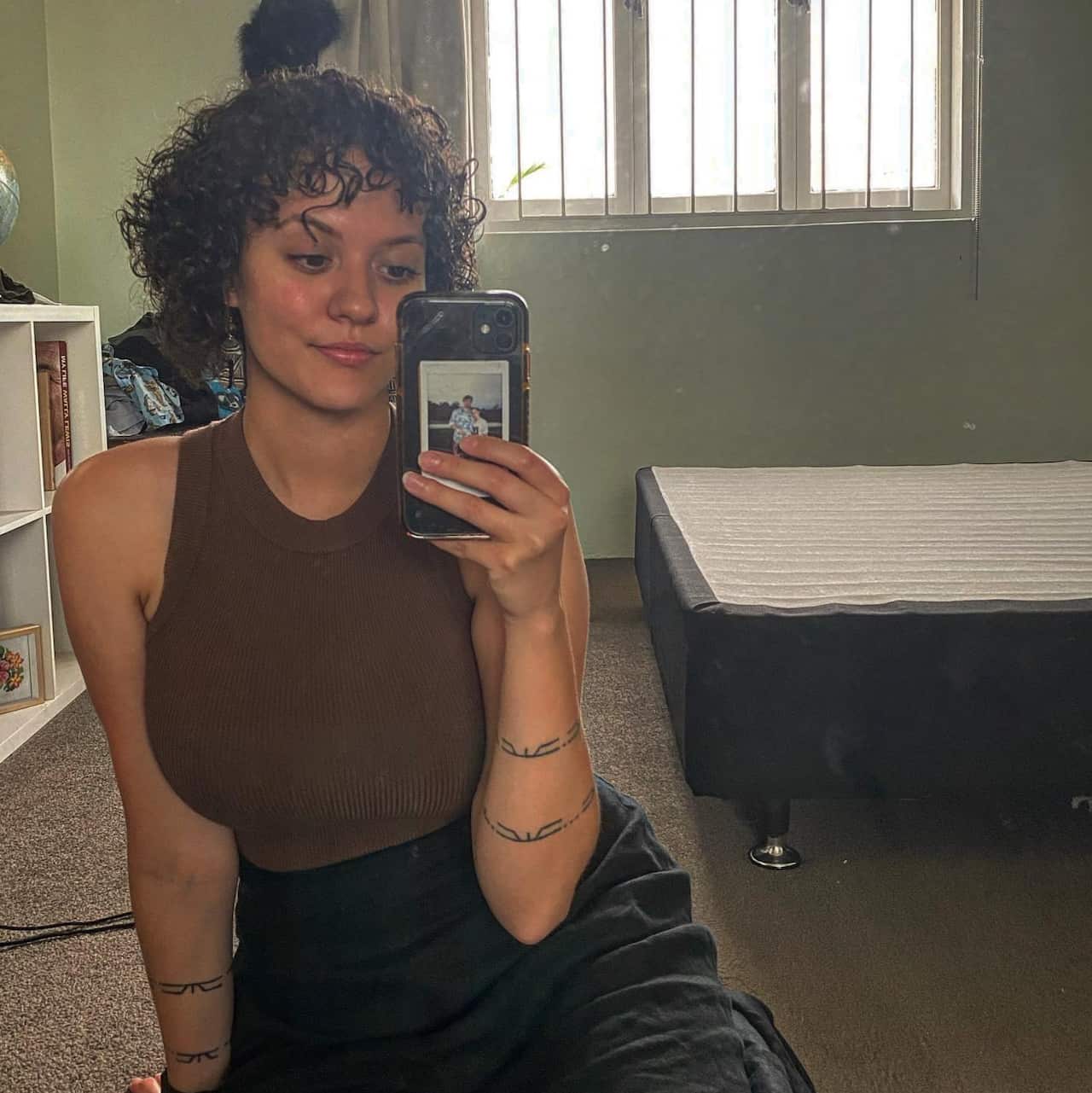 A woman sitting on the floor in front of a bed taking a selfie in the mirror