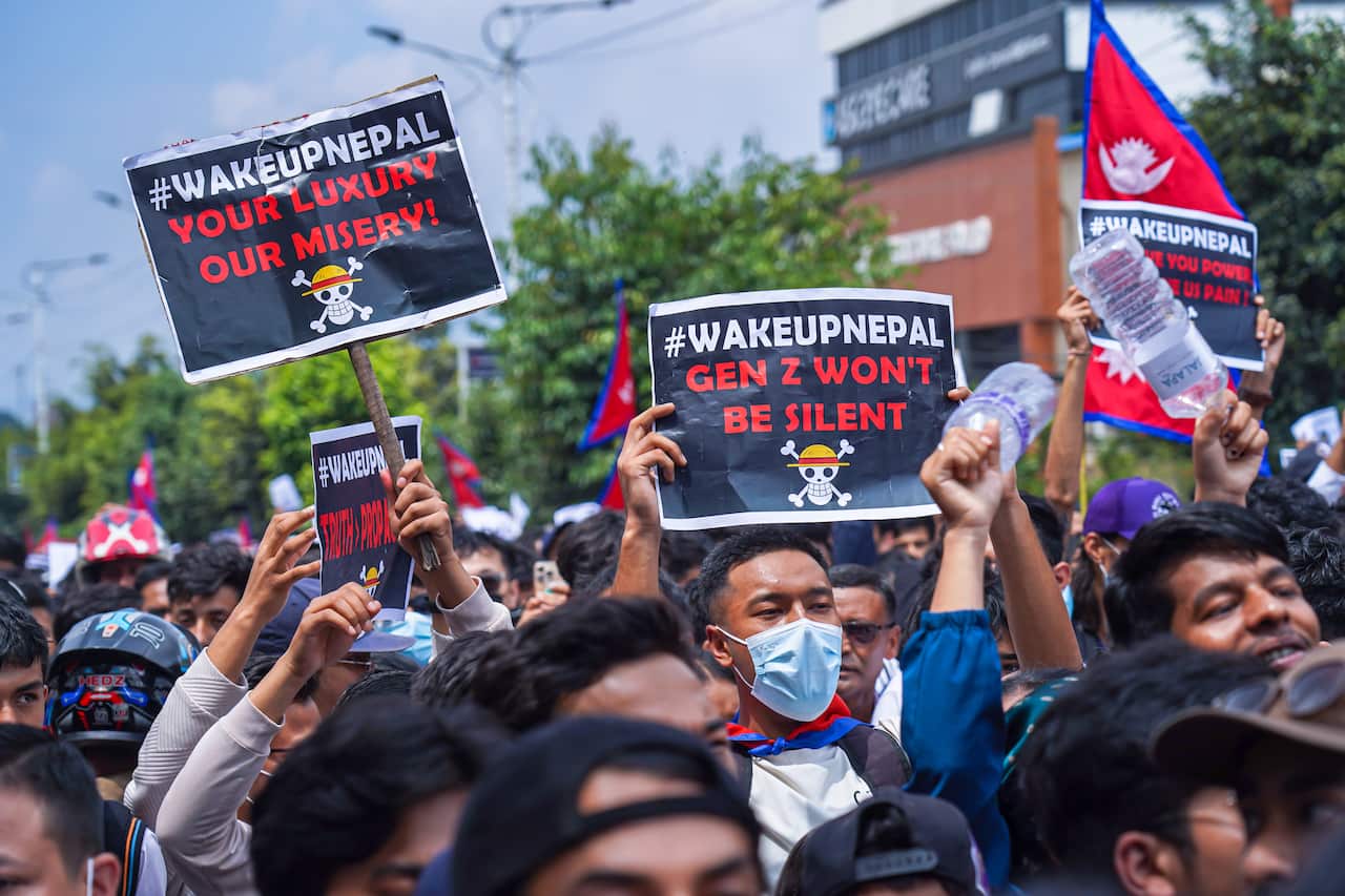 A group of people hold up signs which read '#WakeUpNepal Your Luxury, Our Misery' and '#WakeUpNepal Gen Z Won't Be Silent'. The signs have a skull and crossbones icon. Some people also hold Nepali flags. 
