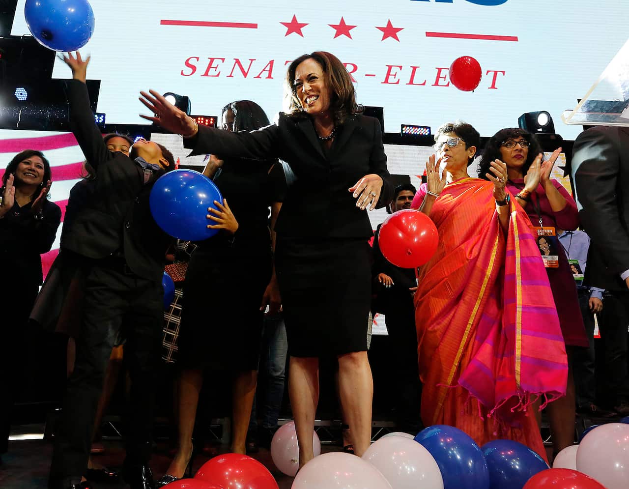 Kamala Harris stands on stage, waving to an unseen crowd. Behind her, a diverse group of supporters gathers, and a large screen displays the words Senator-elect.