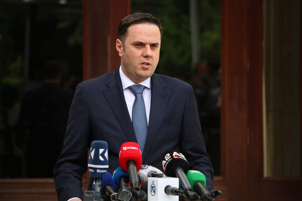 A man in a dark suit and blue patterned tie speaks behind a row of news microphones during an outdoor press conference.