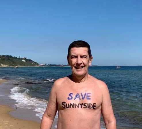 A nude man stands on a beach. Waves and a love heart are drawn on his back, along with the words "Sunnyside Beach!".