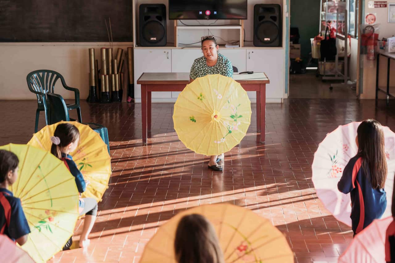 A woman standing at the front of a room and holding an open yellow parasol. Children holding open parasols are facing her.