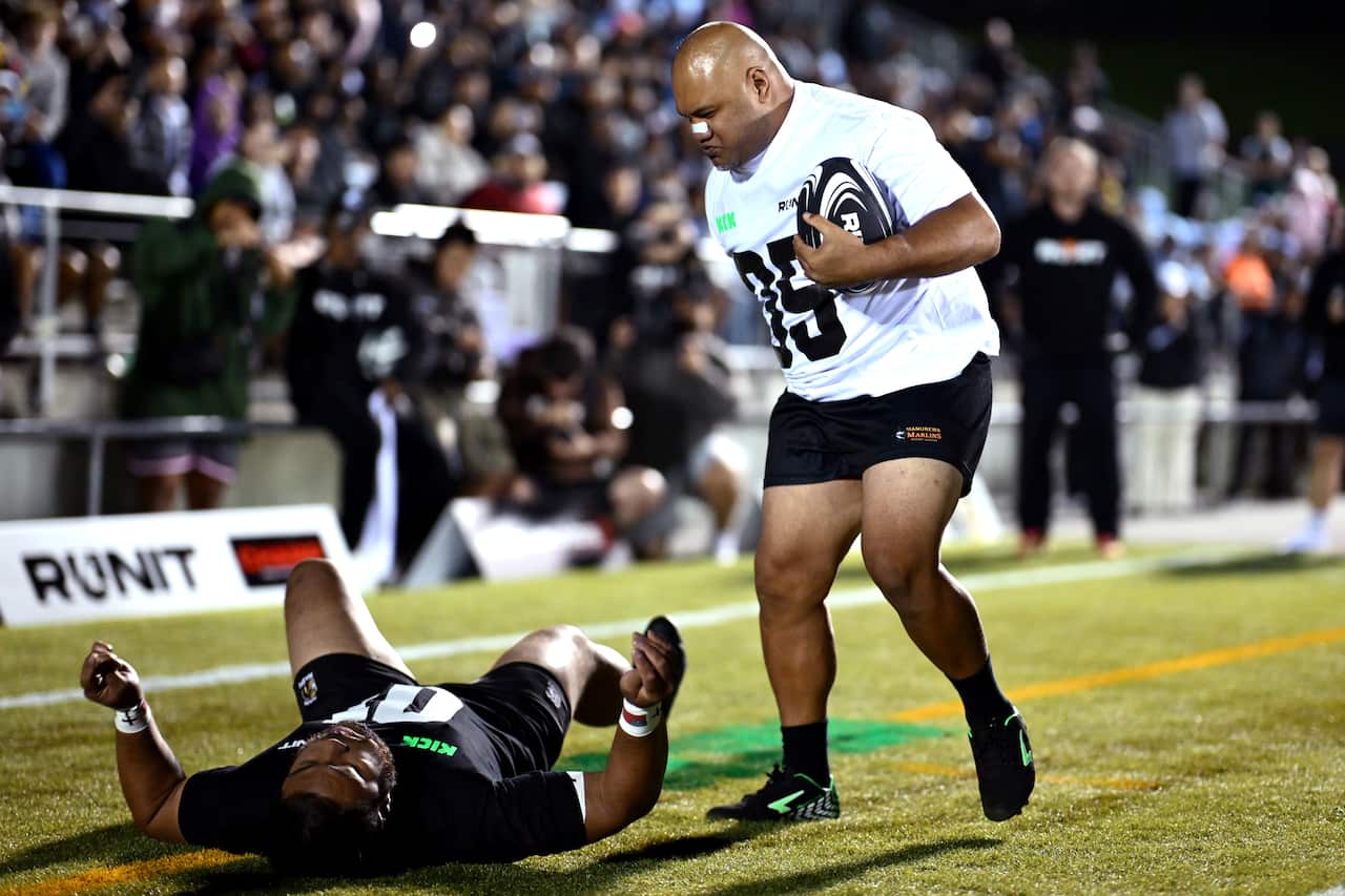 A man standing on a grass field holds a rugby ball while looking down at another man on his back, laying on the grass.