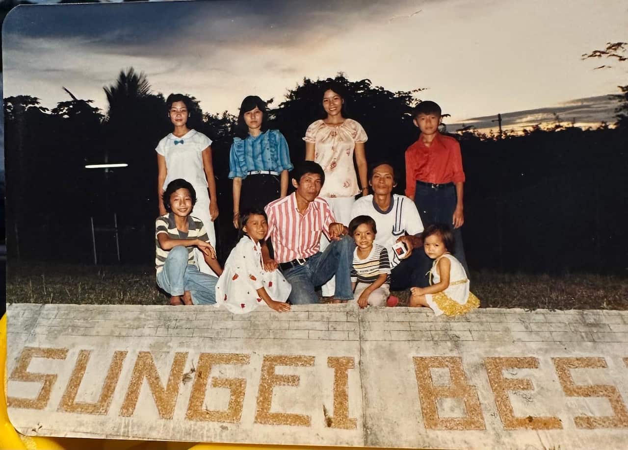 A small group of people, some standing and some kneeling, in front of a large sign that reads Sungei Besi