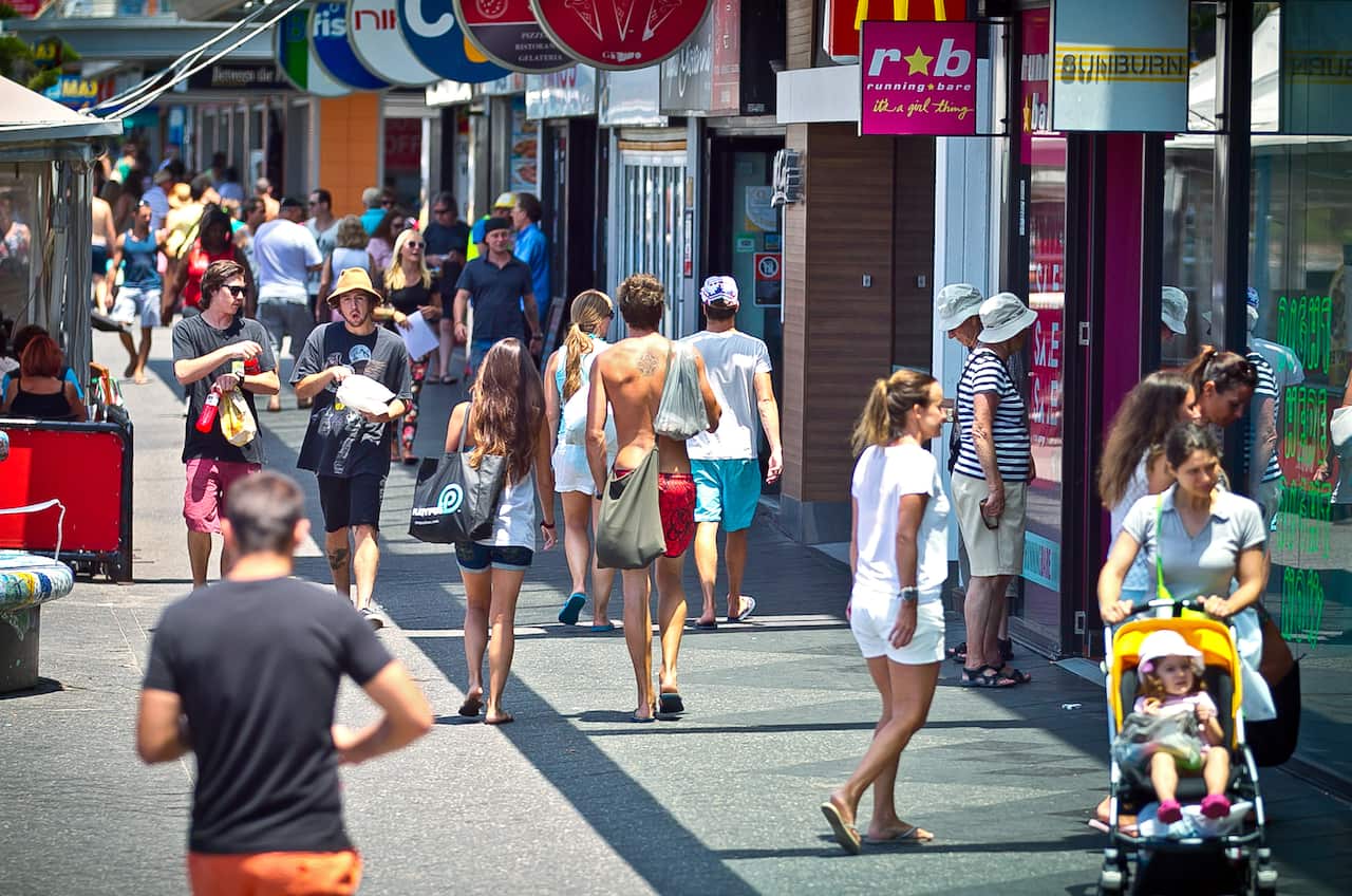 A crowd of people in summer attire strolls down a sun-drenched outdoor shopping promenade lined with various storefronts and colorful overhead signs.
