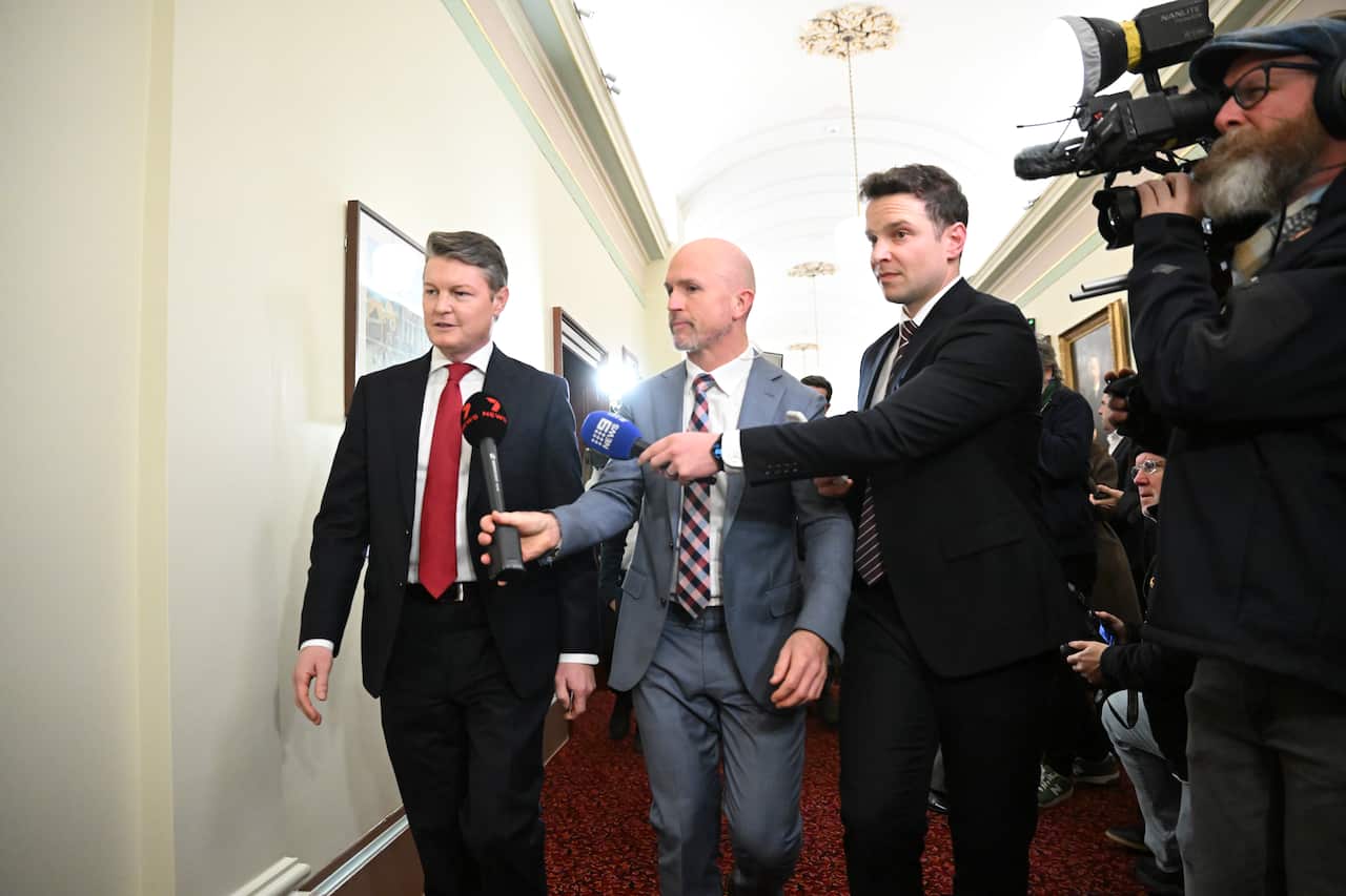 A man in a black suit walking down a corridor. TV reporters with microphones are walking beside him.