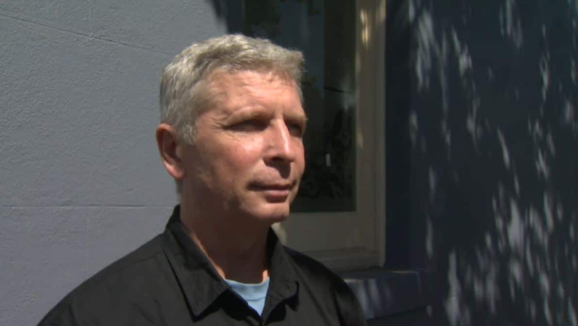 A clean-shaven middle-aged man with grey hair wearing a black polo shirt stares forward with a serious expression on his face. 