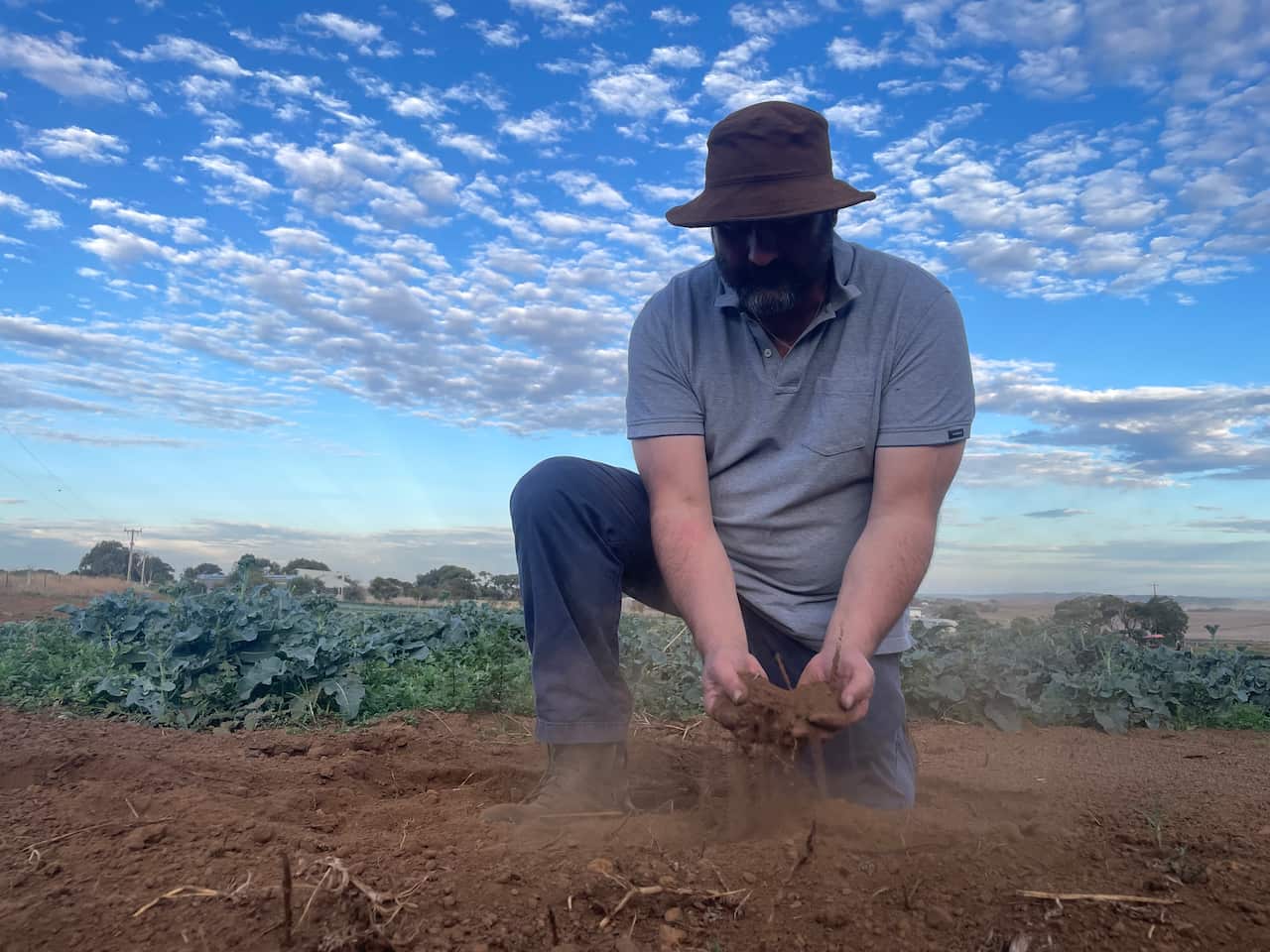 A man wearing jeans and a hat picks up some dirt from the ground.