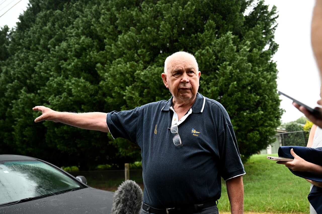 An elderly man is standing outside and speaking to journalists with one hand pointing in his right direction.