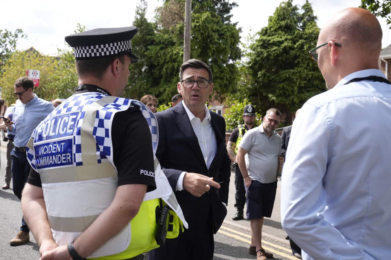 UK PM Andy Burnham in a suit talks to two men in uniform with their backs to the camera.