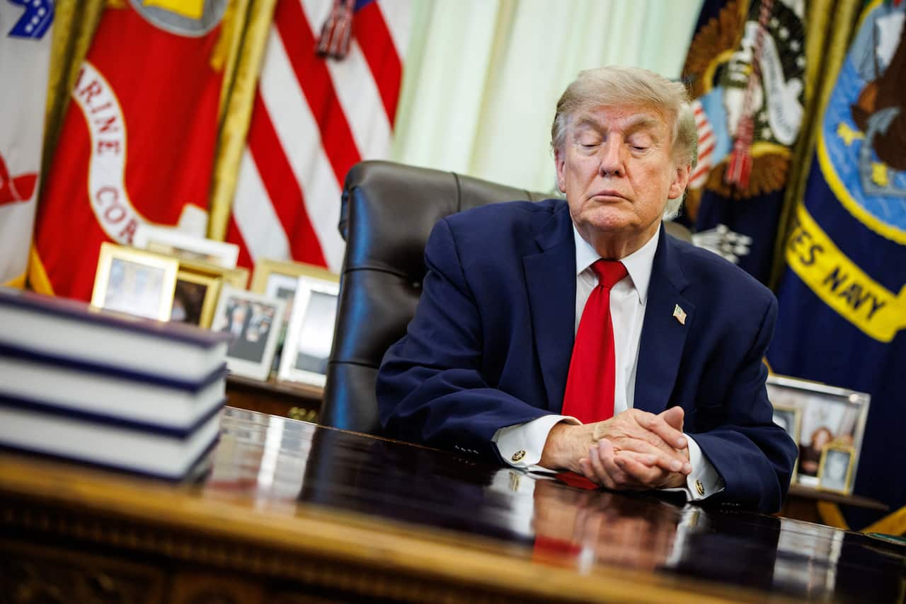 Donald Trump, wearing a blue suit and red tie, sits at his desk clasping his hands together.