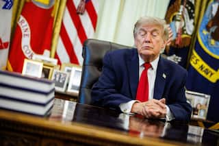 Donald Trump, wearing a blue suit and red tie, sits at his desk clasping his hands together.