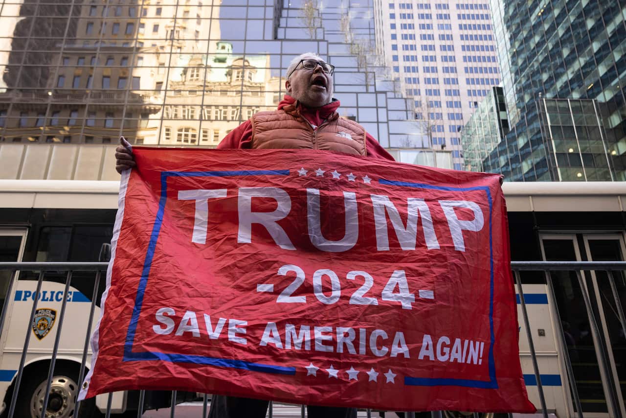 A man stands outside a building holding a large red and blue banner which reads: Trump - 2024 - Save America Again!