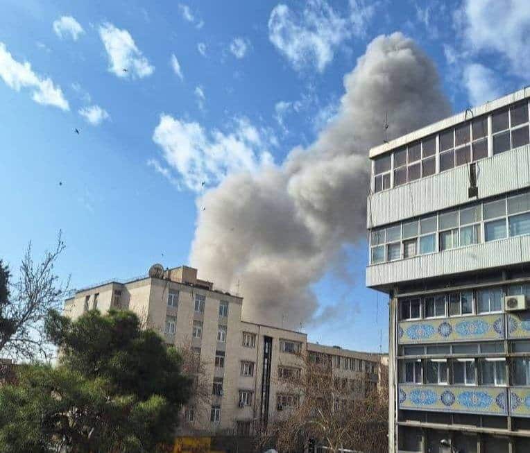 A thick plume of grey smoke rises into a blue sky from behind urban apartment buildings.