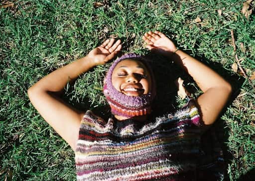 A woman wearing a multicoloured knitted balaclava and a top is smiling as she lies on the grass.