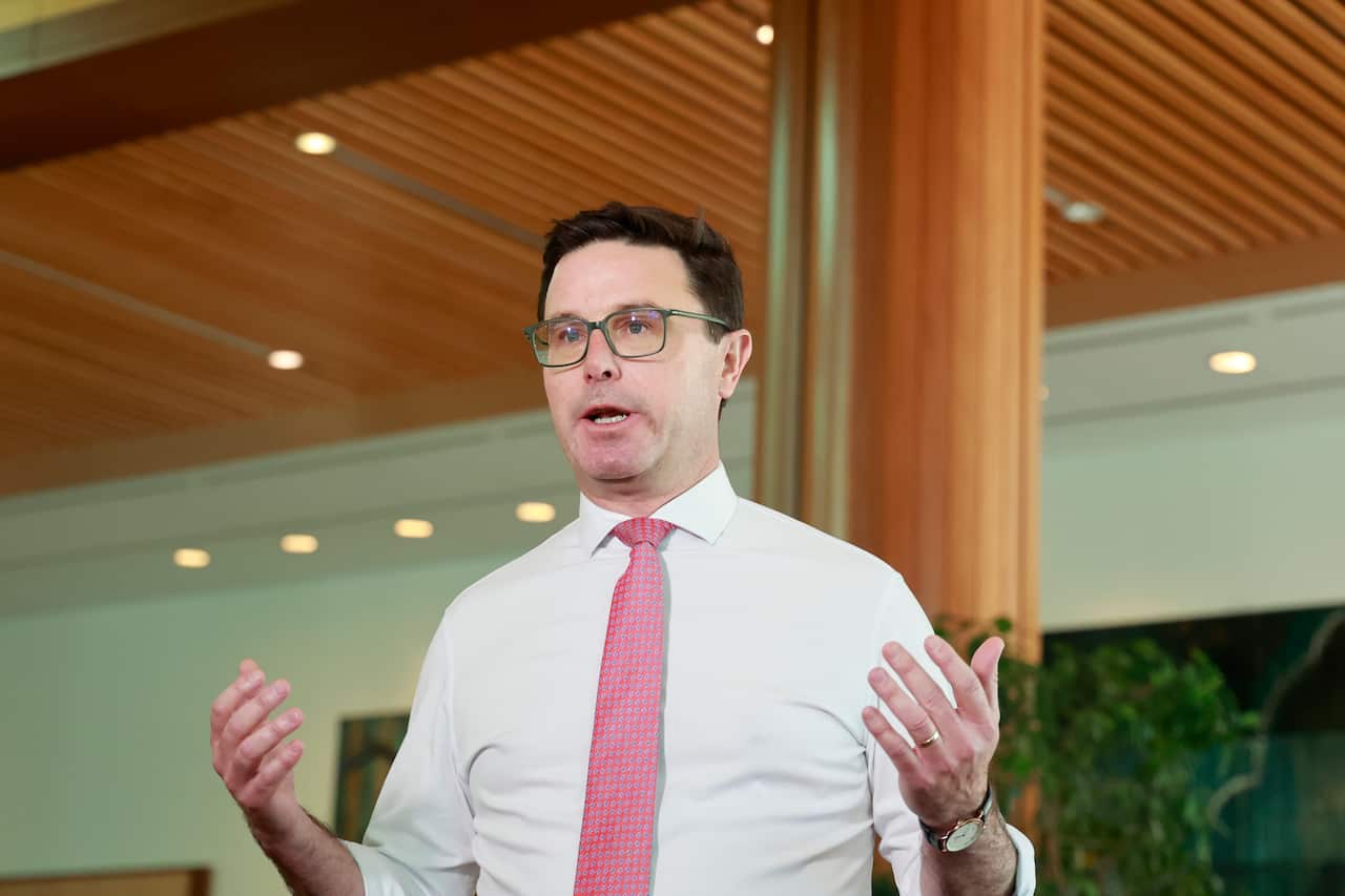 A middle-aged white man wearing a white shirt, red tie and glasses. His hands are slightly raised beside him as he speaks at a press conference. 