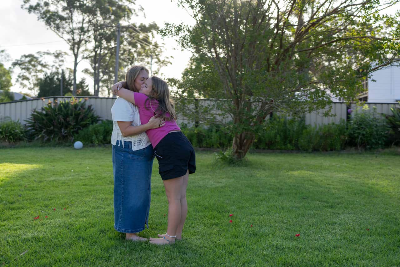 A middle-aged woman wearing a white shirt and denim skirt hugs a young girl who is wearing a pink top and black shorts while standing in a garden.