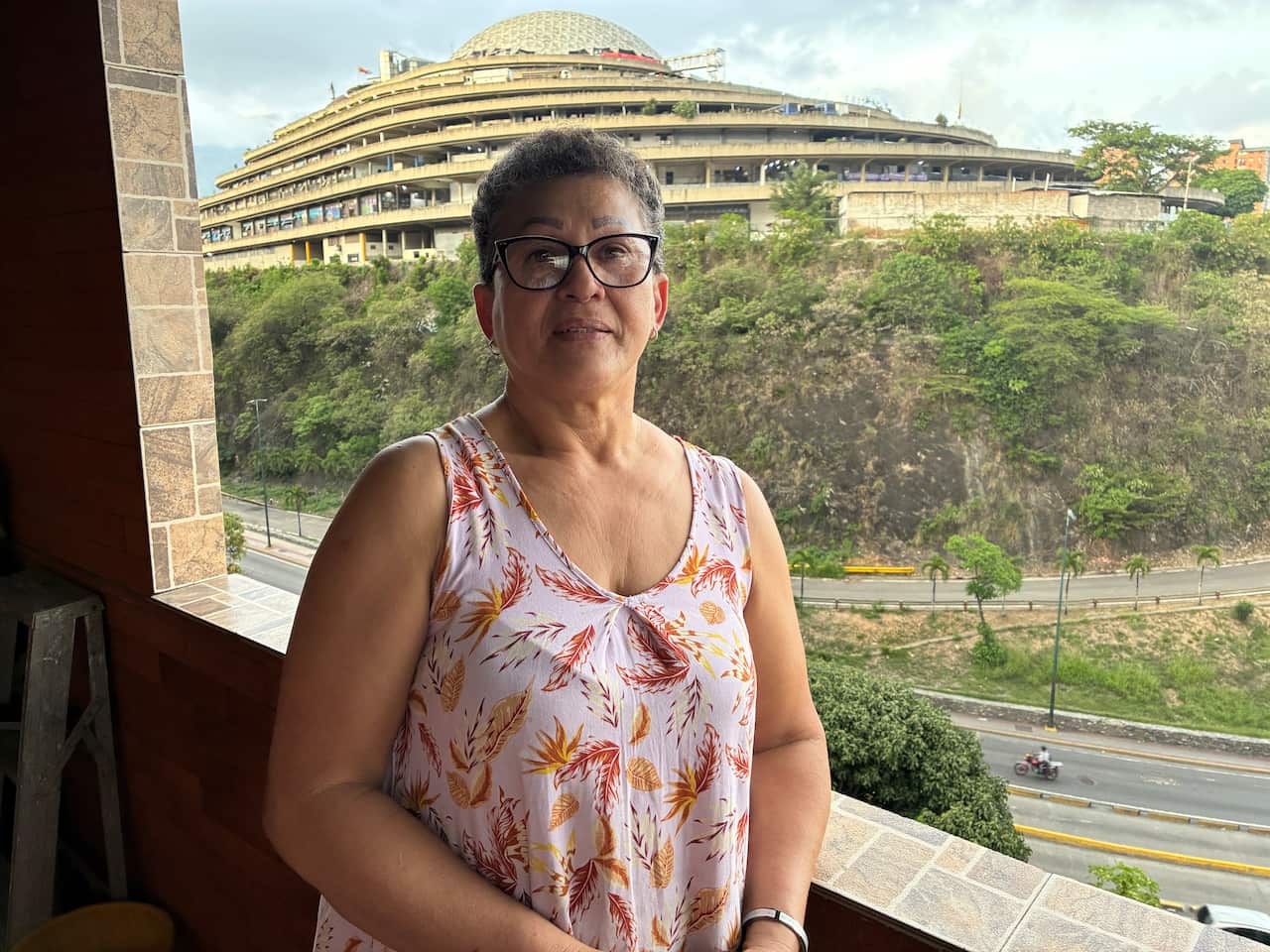A middle-aged woman in a floral dress stands in front of a balcony looking over a road and a large building.