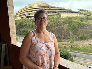 A middle-aged woman in a floral dress stands in front of a balcony looking over a road and a large building.