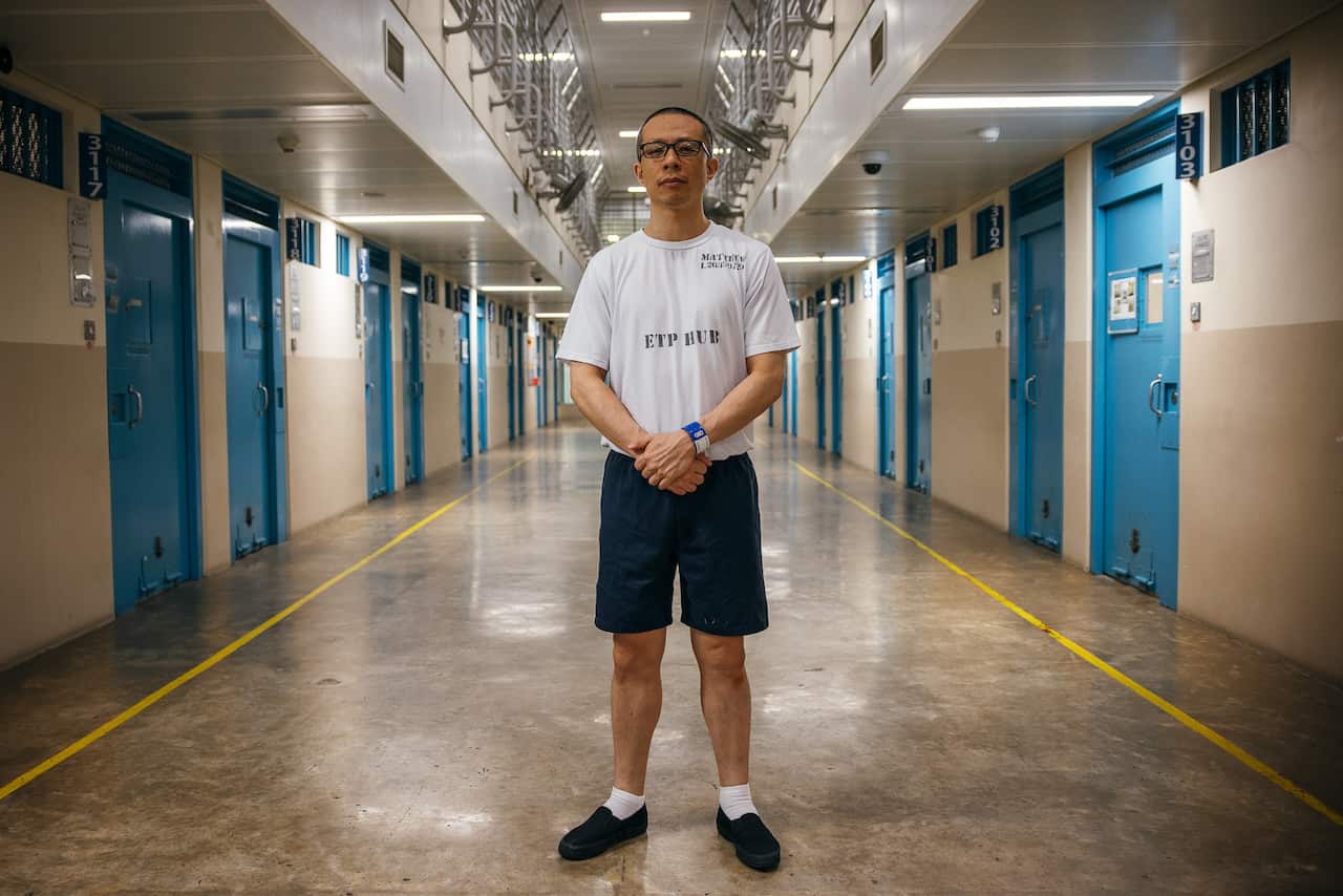 A man wearing glasses, a white t-shirt and black shorts looks straight at the camera. Behind him are the blue doors of prison cells