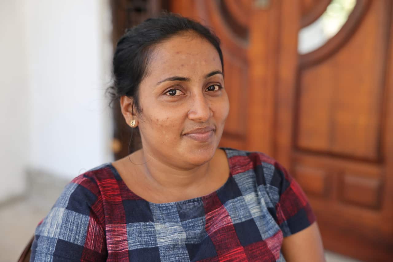 A woman in a checked dress sitting outside a wooden door stares into the camera