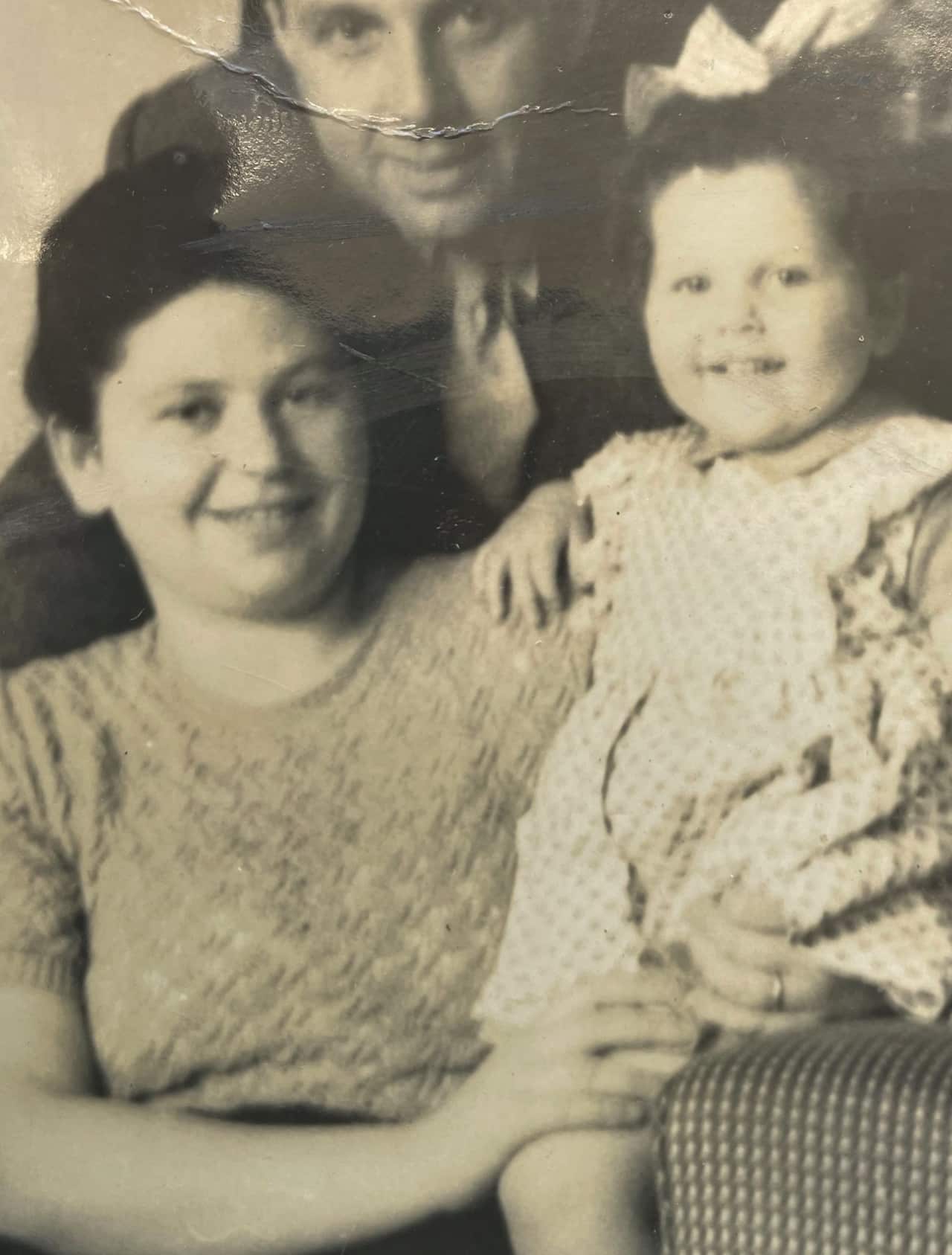 A woman holds a small child on an armchair while a man stands in the background.