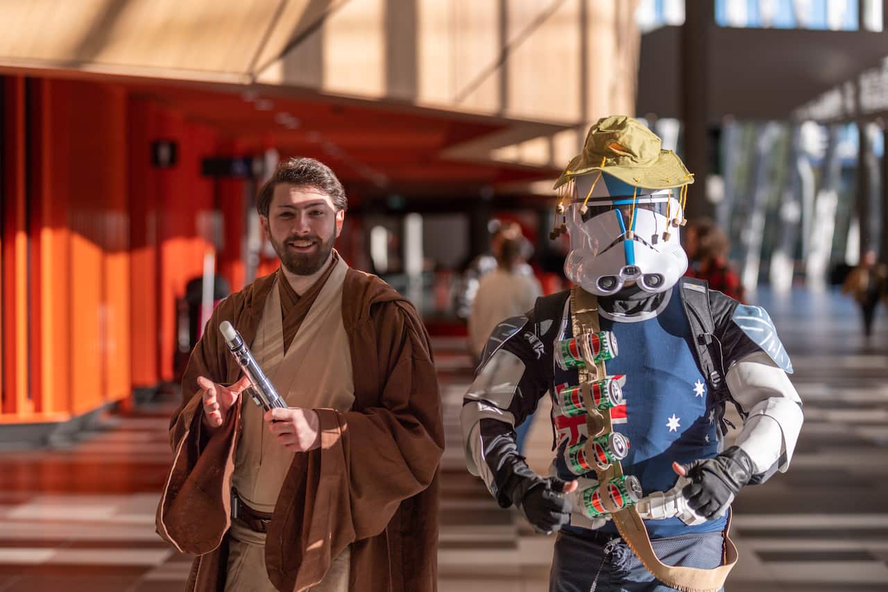 Two people, one man dressed up in brown costume robes, and another wearing a 'droid' helmet from Star Wars, an Australian flag singlet and an 'ammo' strap made of green beer cans.