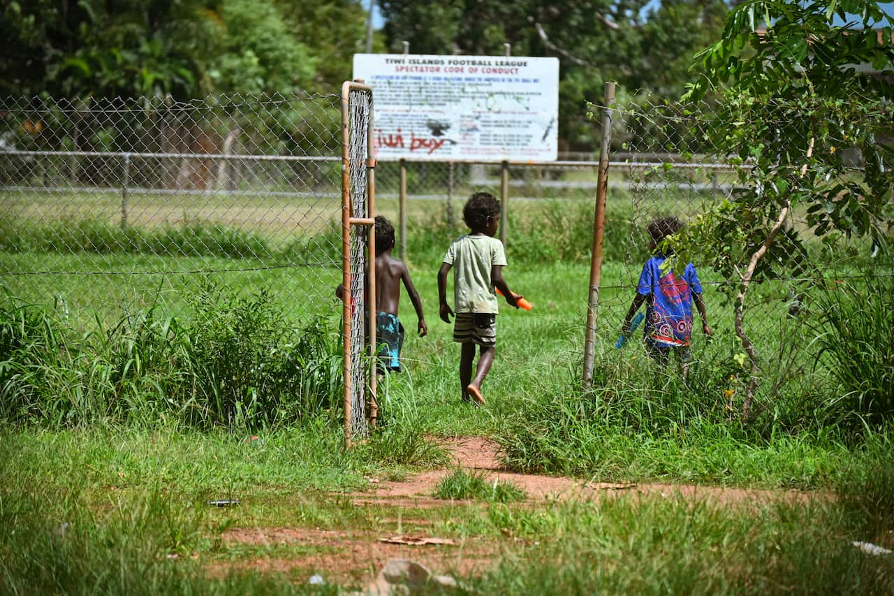Three small children walk away from the camera . They are walking towards a grassy area on the other side of an open gate. A large sign in the distance lists the rules for the football oval.