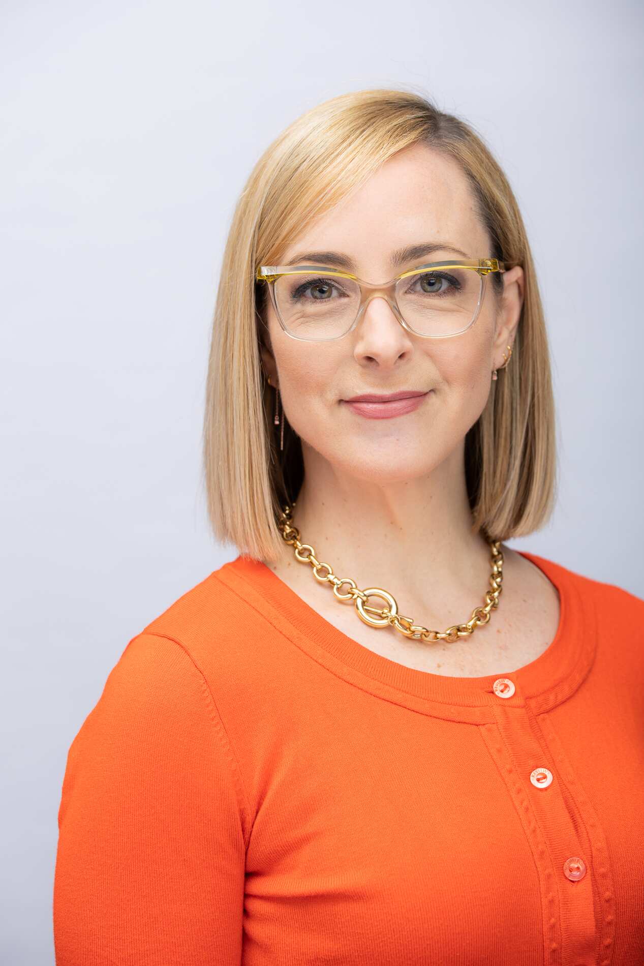 A woman wearing an orange top and glasses looks forward. 