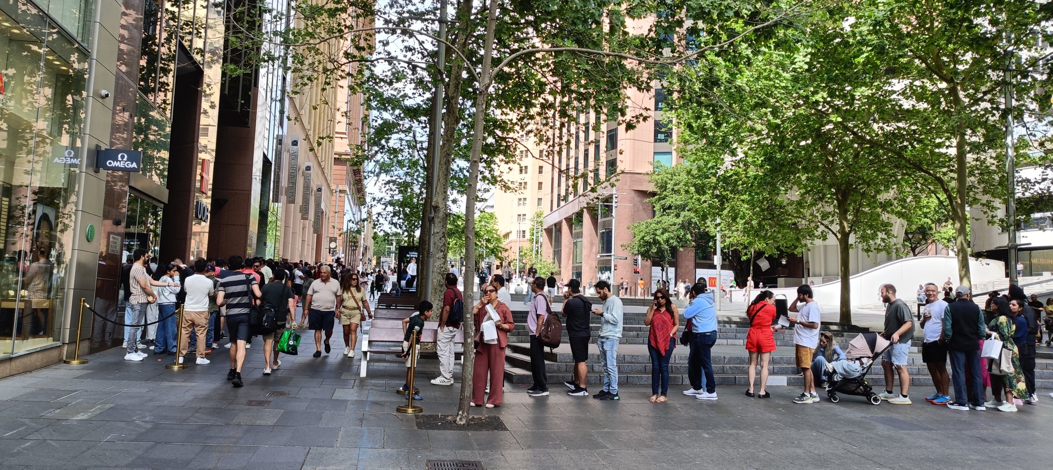 A line of people queue outside a commercial building with an Omega sign, with other pedestrians walking past.