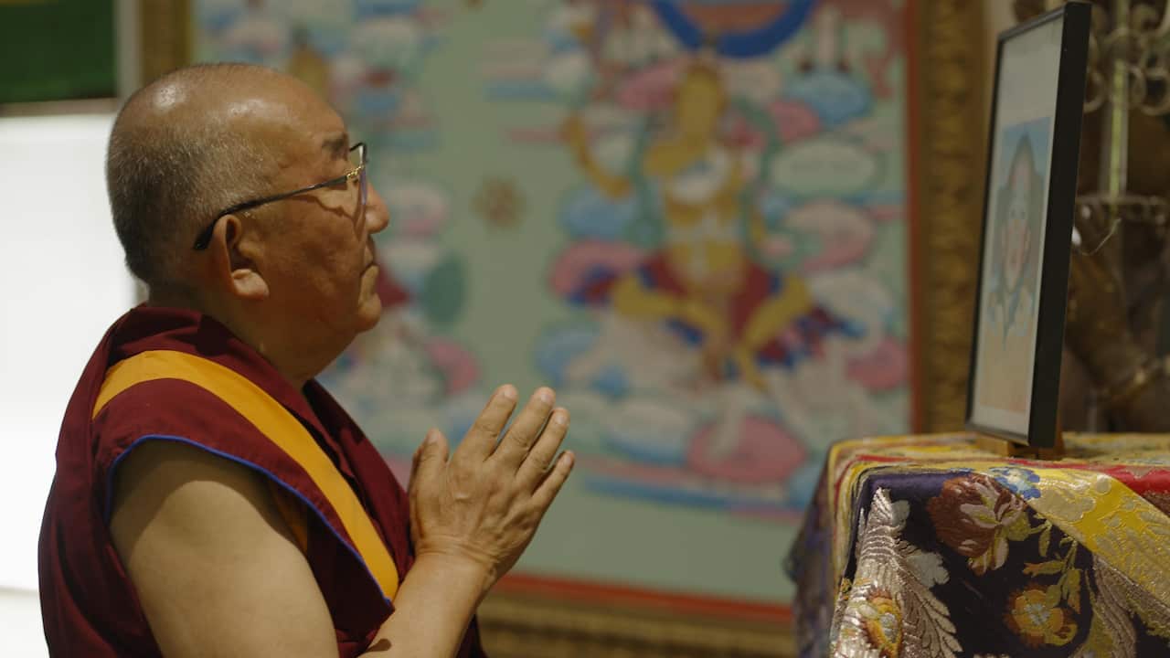 A monk with a shaved head and wearing a maroon and yellow robe faces a framed picture on a table with his hands pressed together in front of him