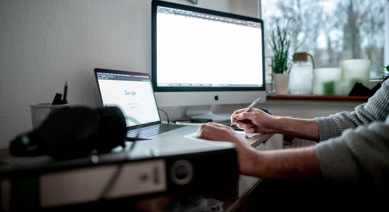 A man at a desk with a computer and laptop screen. Only his arms are visible.