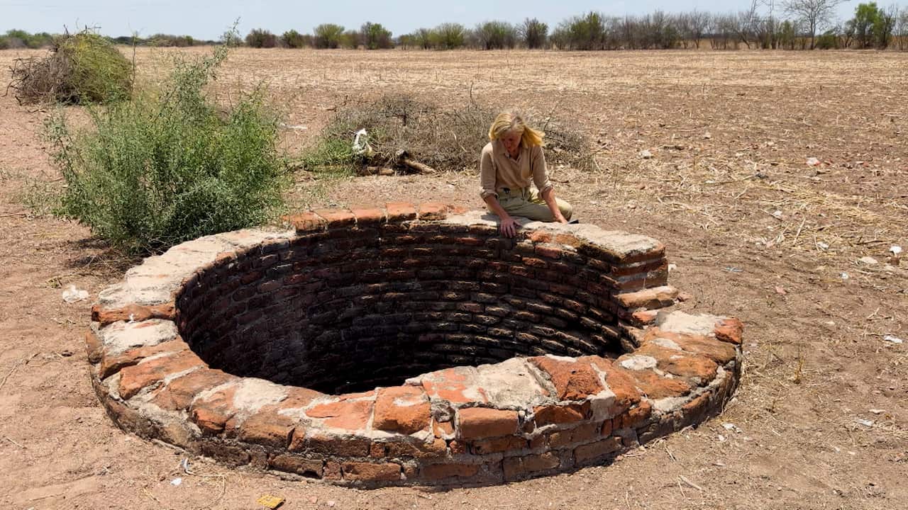 A blonde woman sits on the edge a large well, looking down inside it, in a dry, barren field