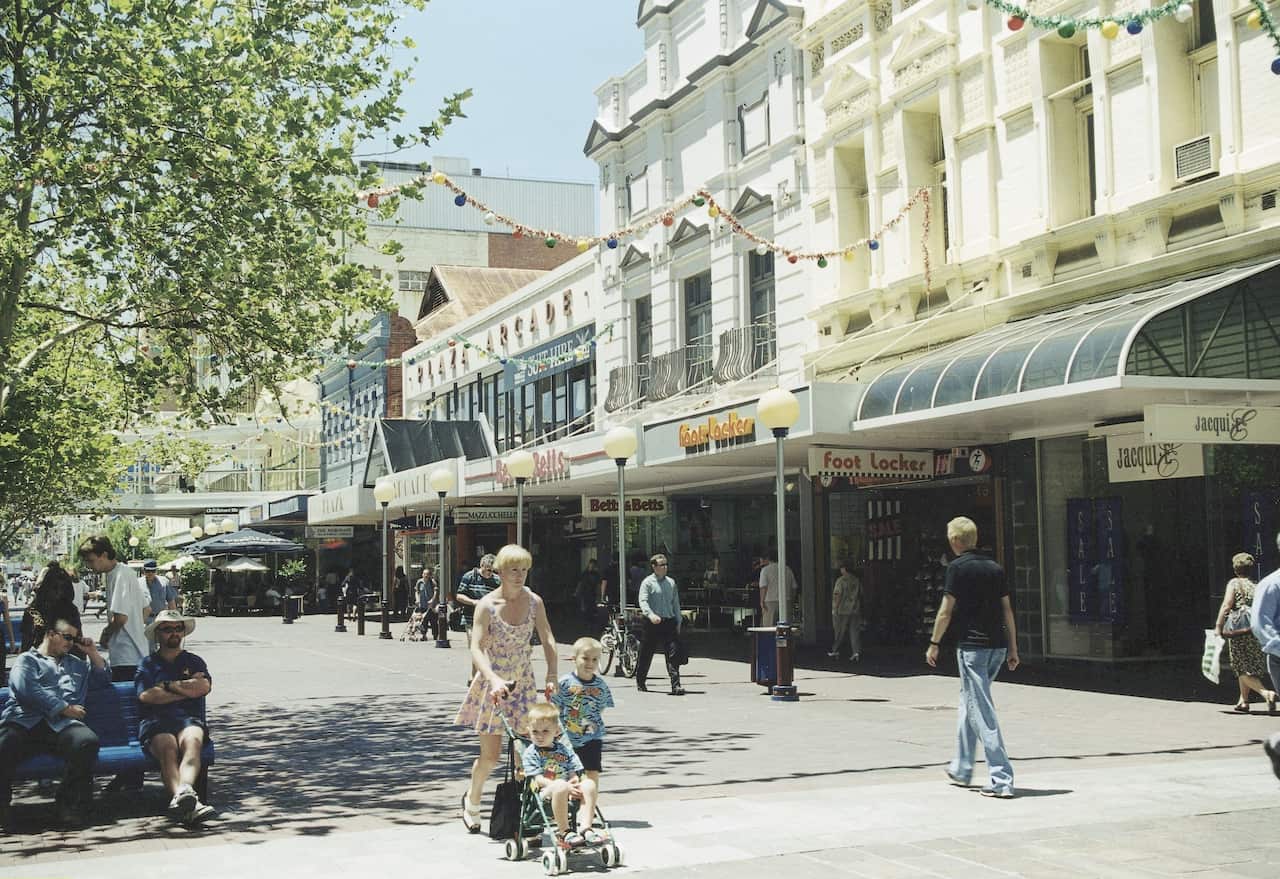 A photo from the 1990s showing a shopping high street with people walking and sitting around. Stores lining the street inclue Jacqui E and Foot Locker