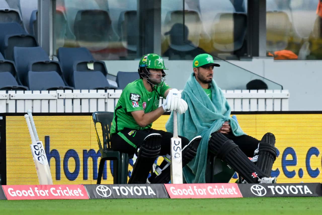 Two cricket batsmen sitting in chairs on the sidelines. One (right) has a large towel over his shoulders. 