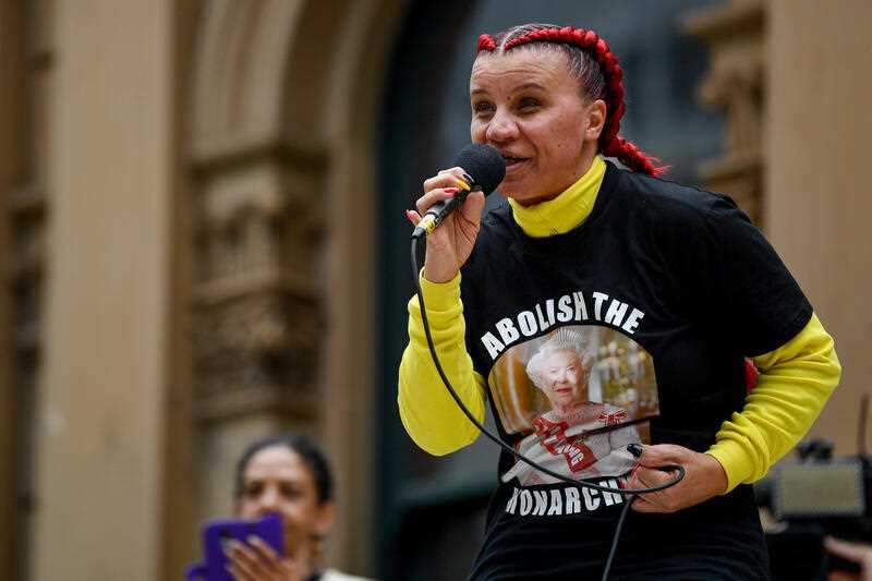 A woman wearing an t-shirt with the words abolish the monarchy speaks into a microphone