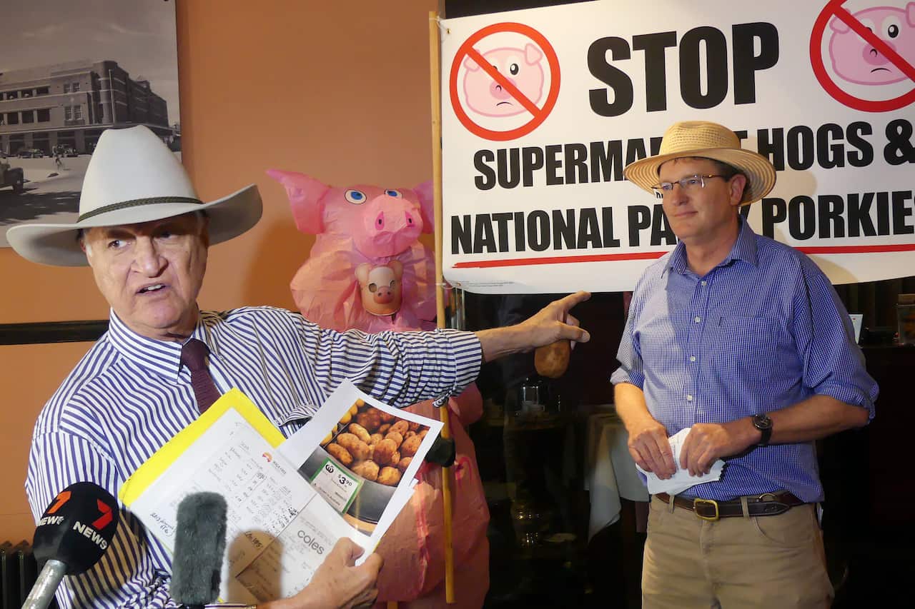 A man speaking in front of microphones while pointing at another man standing behind him.