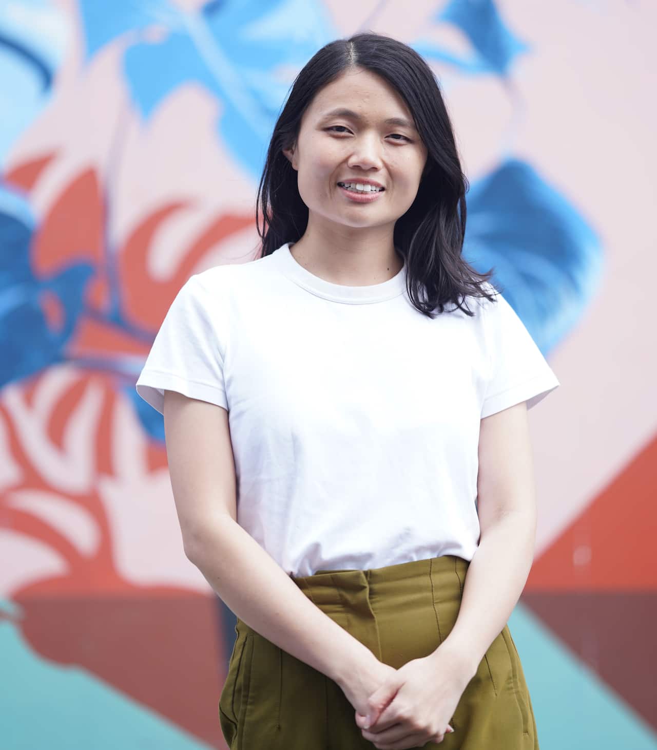 A woman stands with her hands folded in a relaxed manner.